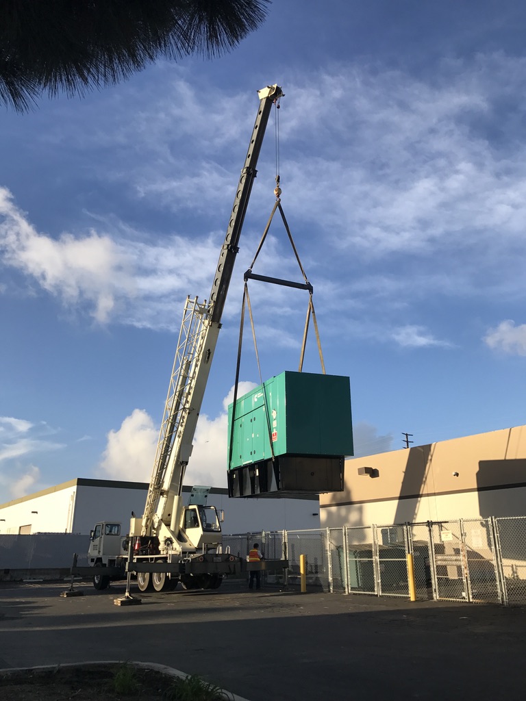 White rig truck lifting a big green generator over a chain-linked warehouse fence