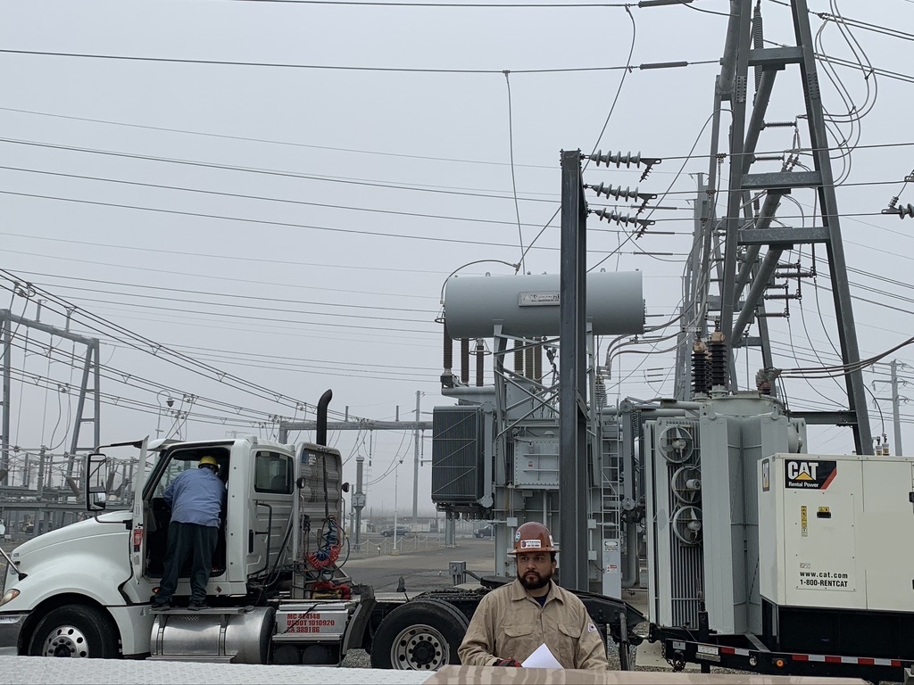 Guy in a beige jacket and red hat standing in front of a truck with a platform attachment containing a white generator