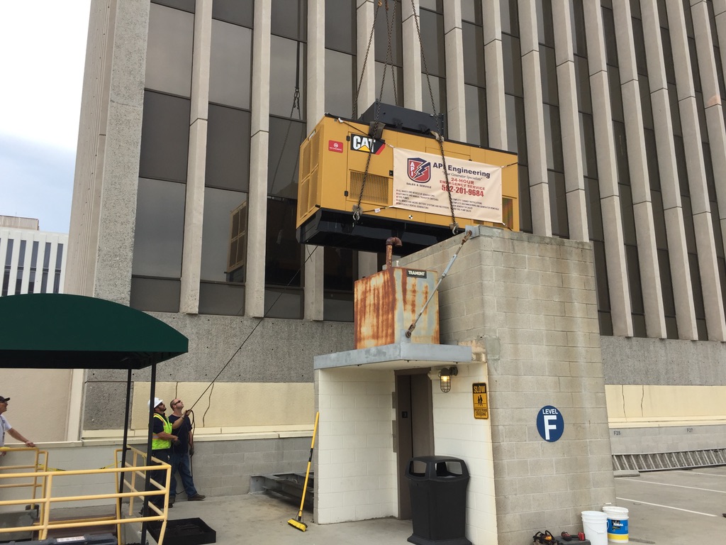 Yellow CAT generator being lifted above a grey, roofless building using black ropes